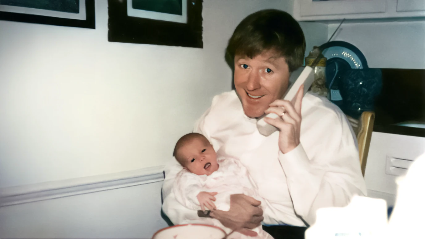 David Booth smiling and holding an infant with one arm and a phone to his ear with his other hand.