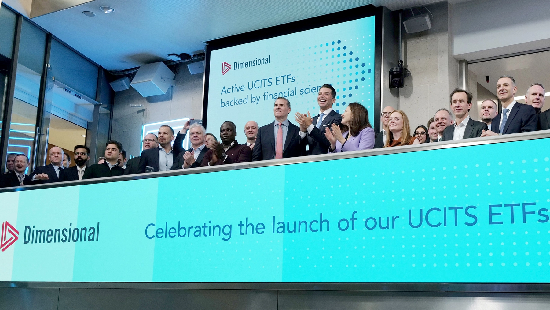 A crowd of people on a balcony at the London Stock Exchange. A screen behind them says “Dimensional active UCITS ETFs backed by financial science.” A digital banner below them says “Dimensional celebrating the launch of our UCITS ETFs.”